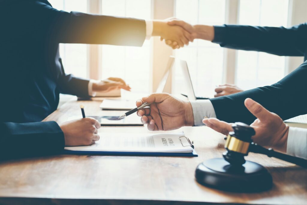 Two people in business attire shaking hands across a desk during a legal meeting, with a signed document on a clipboard, a pen being exchanged, and a judge’s gavel visible in the foreground, suggesting a finalized agreement or settlement in a professional office setting.
