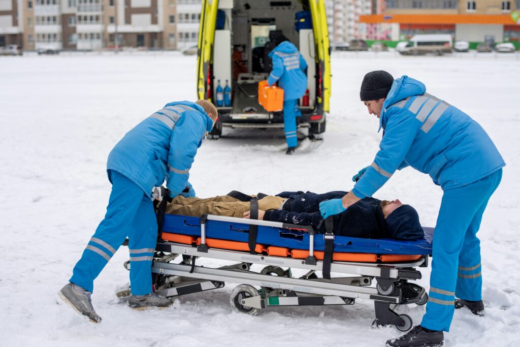 Emergency medical responders in blue winter uniforms securing an injured person onto a stretcher in a snowy outdoor area, with an ambulance parked behind them and one responder retrieving medical equipment from the open rear doors while two others stabilize the patient for transport.