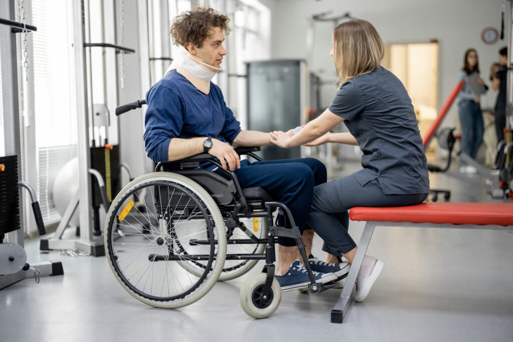 A physical therapist seated on a red therapy bench assists a young man in a wheelchair during a rehabilitation session, supporting his forearm and wrist while he wears a cervical neck brace, with exercise machines and other patients visible in a bright clinical physical therapy gym.