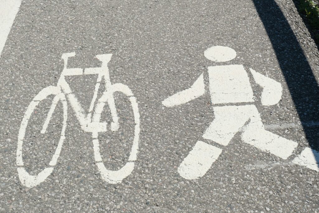 White painted pavement symbols show a simple outline of a bicycle on the left and a walking pedestrian on the right, stenciled onto gray asphalt with visible stone texture, indicating a shared path or designated area for cyclists and pedestrians, viewed from above in bright daylight with sharp shadows along the edge of the road.
