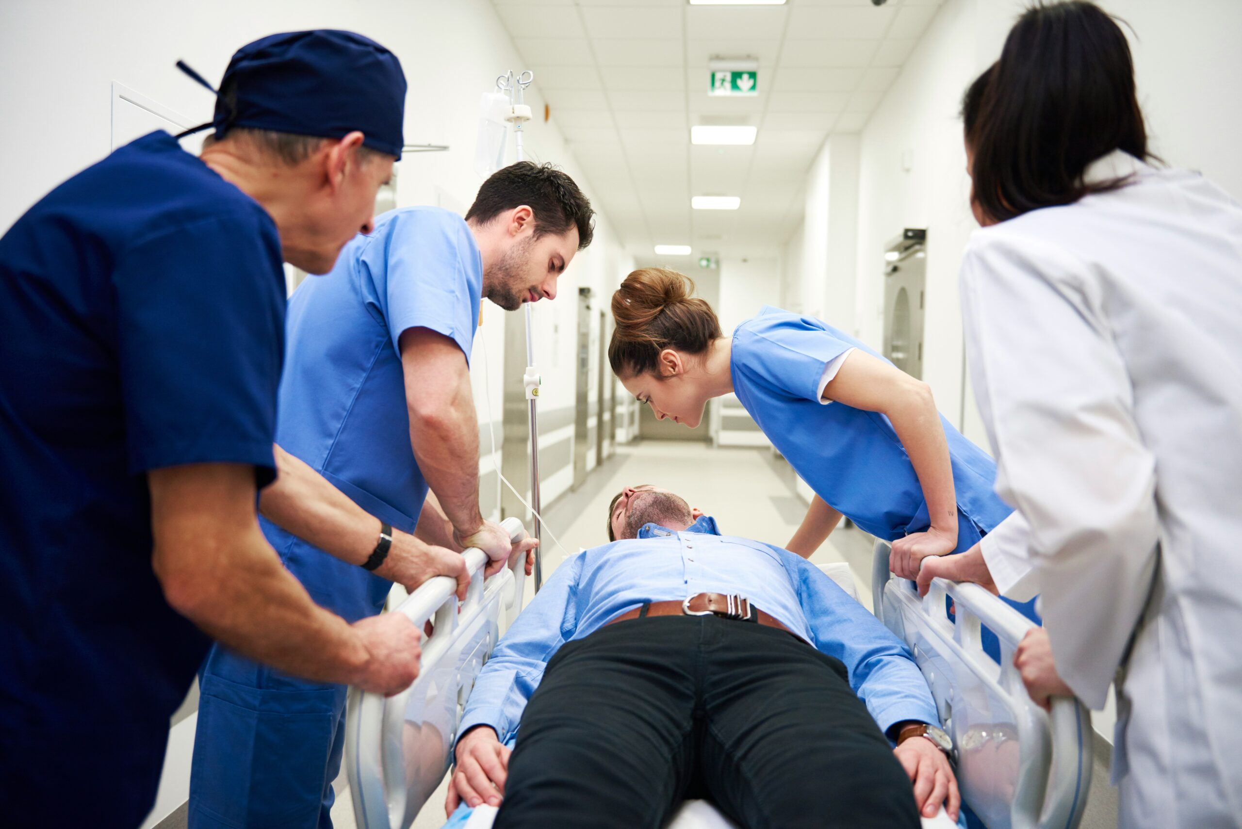 Hospital staff in blue scrubs and white coats urgently push a patient lying on a hospital gurney down a bright medical corridor, with an IV stand attached to the bed, one clinician leaning over to check the patient while others guide the rails, conveying an emergency response and coordinated medical care in a clinical hallway setting.