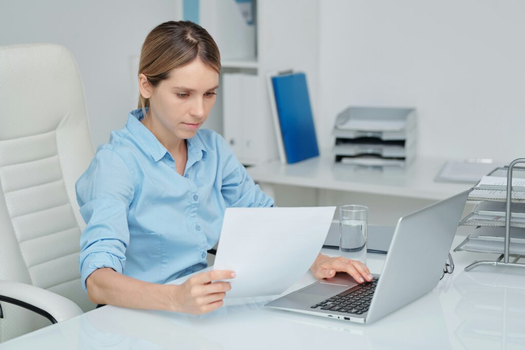Woman seated at a clean white office desk, wearing a light blue button-up shirt, focused on reviewing a printed document while using a laptop, with her left hand holding the paper and her right hand on the keyboard, a glass of water beside the laptop, stacked document trays and folders in the background, and a minimal, bright workspace suggesting administrative or professional paperwork review.