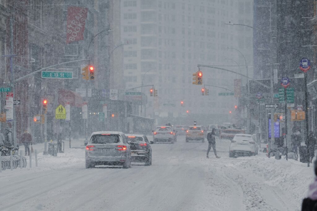 Heavy snowfall blankets a wide city street lined with tall buildings, reducing visibility as cars move slowly through slushy lanes under glowing red traffic lights, while a pedestrian carefully crosses the road amid falling snow, snowbanks piled along the sidewalks, and street signs and signals partially obscured by the storm.