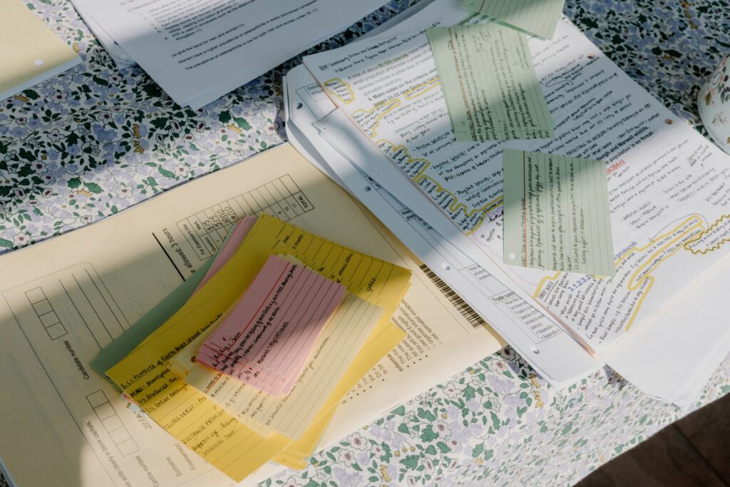 Stack of printed documents and paperwork spread across a table with handwritten notes, highlighted sections, and multiple colored index cards containing handwritten text placed on top, suggesting organization and review of detailed information.