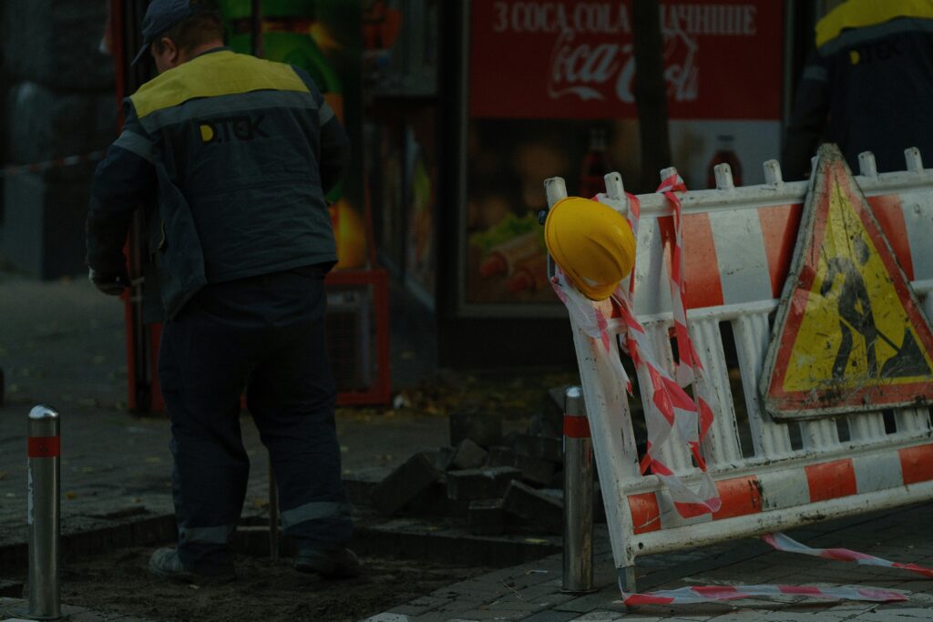Construction worker standing near a street excavation area beside a red-and-white safety barrier with warning tape and a yellow hard hat hanging on it, with a roadwork warning sign visible in the foreground and storefronts blurred in the background.