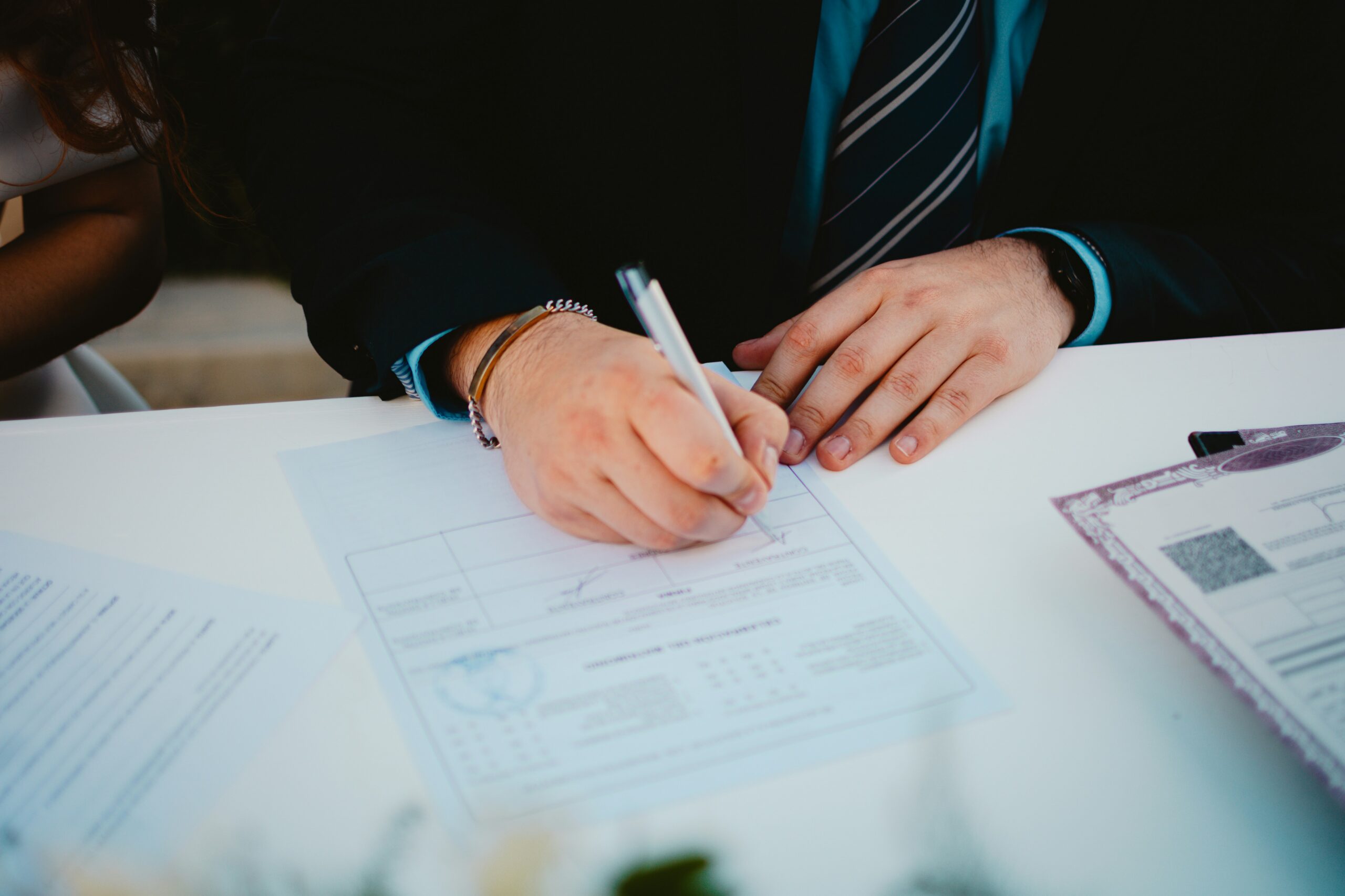 A man in a suit and tie is signing paperwork at a desk, focused on the documents in front of him.