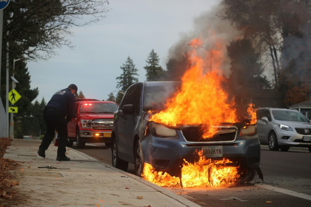 A man stands beside a car engulfed in flames, observing the fire with a concerned expression.