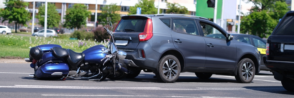 A blue motorcycle lies on its side in the road after colliding with the rear of a gray SUV.