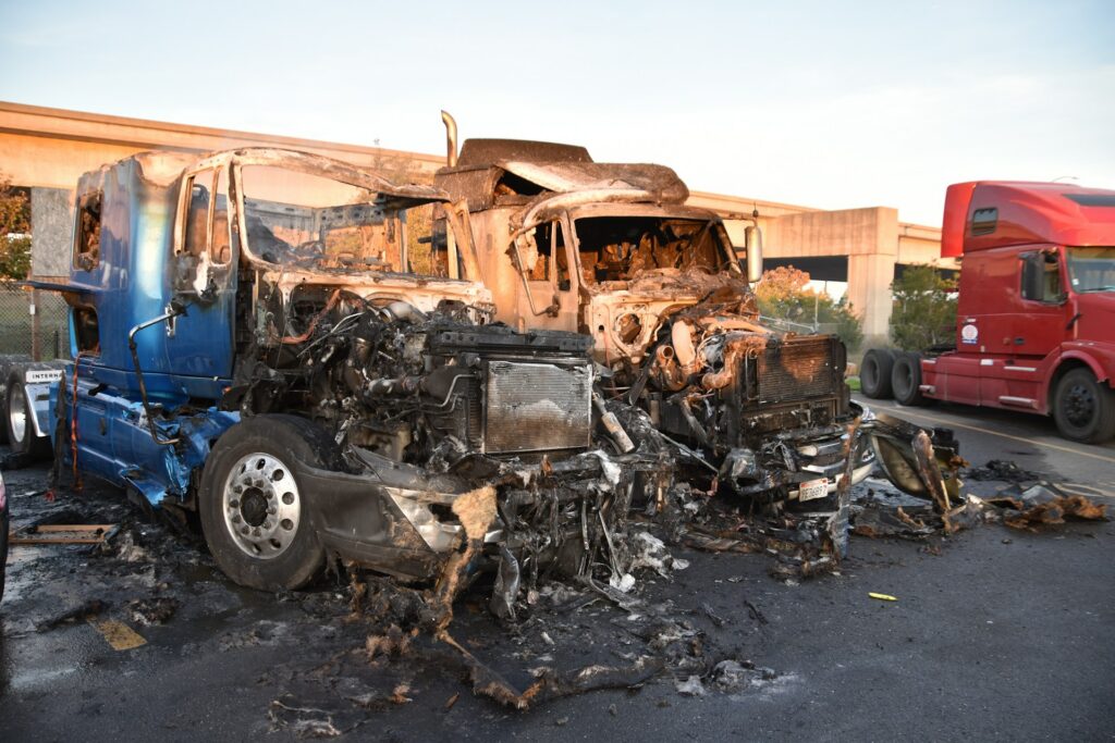 Two burned-out semi-truck cabs sit gutted and charred on a roadside after a fire, with an intact red truck nearby.