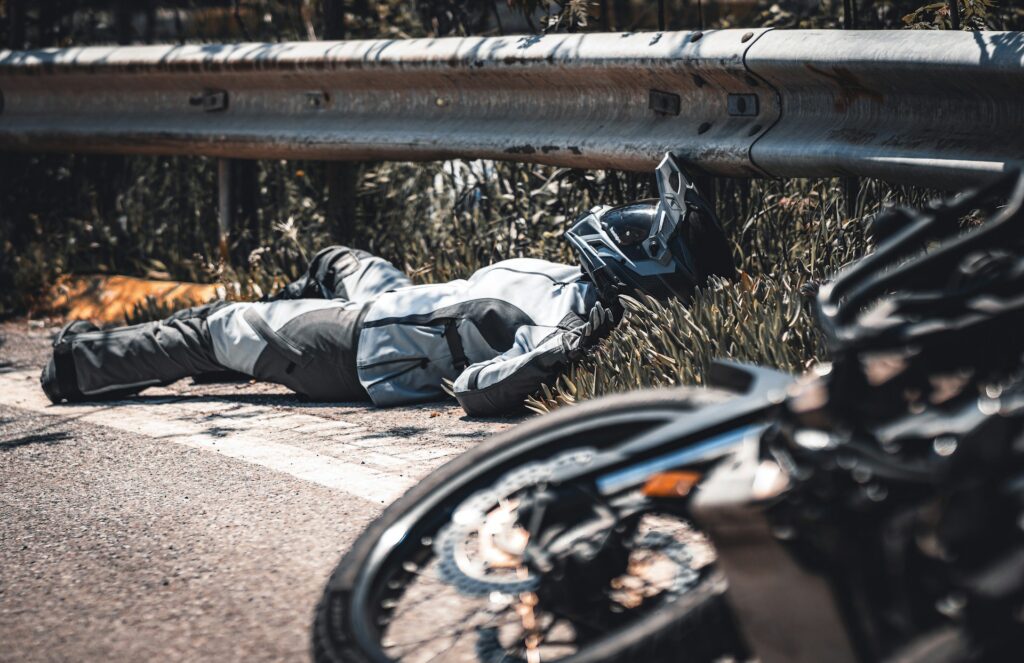 A motorcyclist in protective gear lies motionless on the roadside near a guardrail, helmet nearby, with a motorcycle in the foreground.