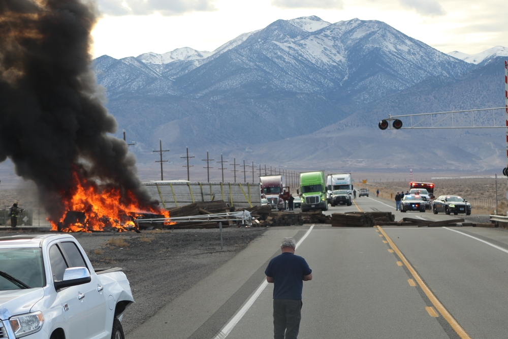 A large truck fire sends black smoke into the air on a highway as police and a bystander look on near snow-capped mountains.